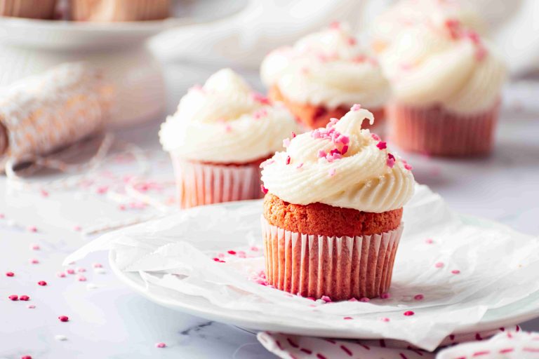 A close-up of a Valentine's Day cupcake with white frosting and pink sprinkles on a plate, with more decorated Valentine's Day cupcakes blurred in the background on a light, airy setting.