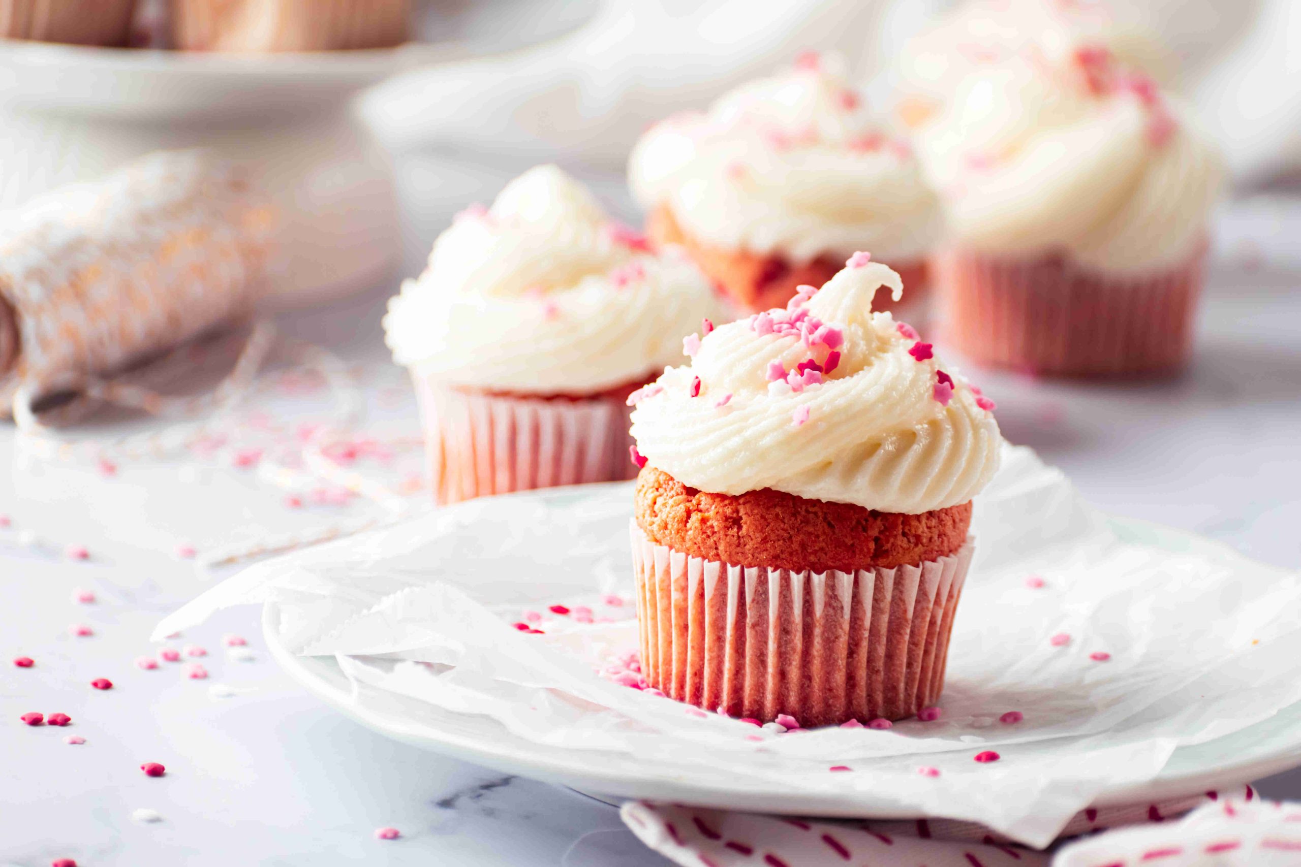 A close-up of a Valentine's Day cupcake with white frosting and pink sprinkles on a plate, with more decorated Valentine's Day cupcakes blurred in the background on a light, airy setting.