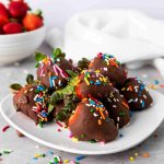 A plate of Valentine's Day Chocolate Covered Strawberries topped with colorful sprinkles sits on a light surface, with a bowl of fresh strawberries and scattered sprinkles in the background.