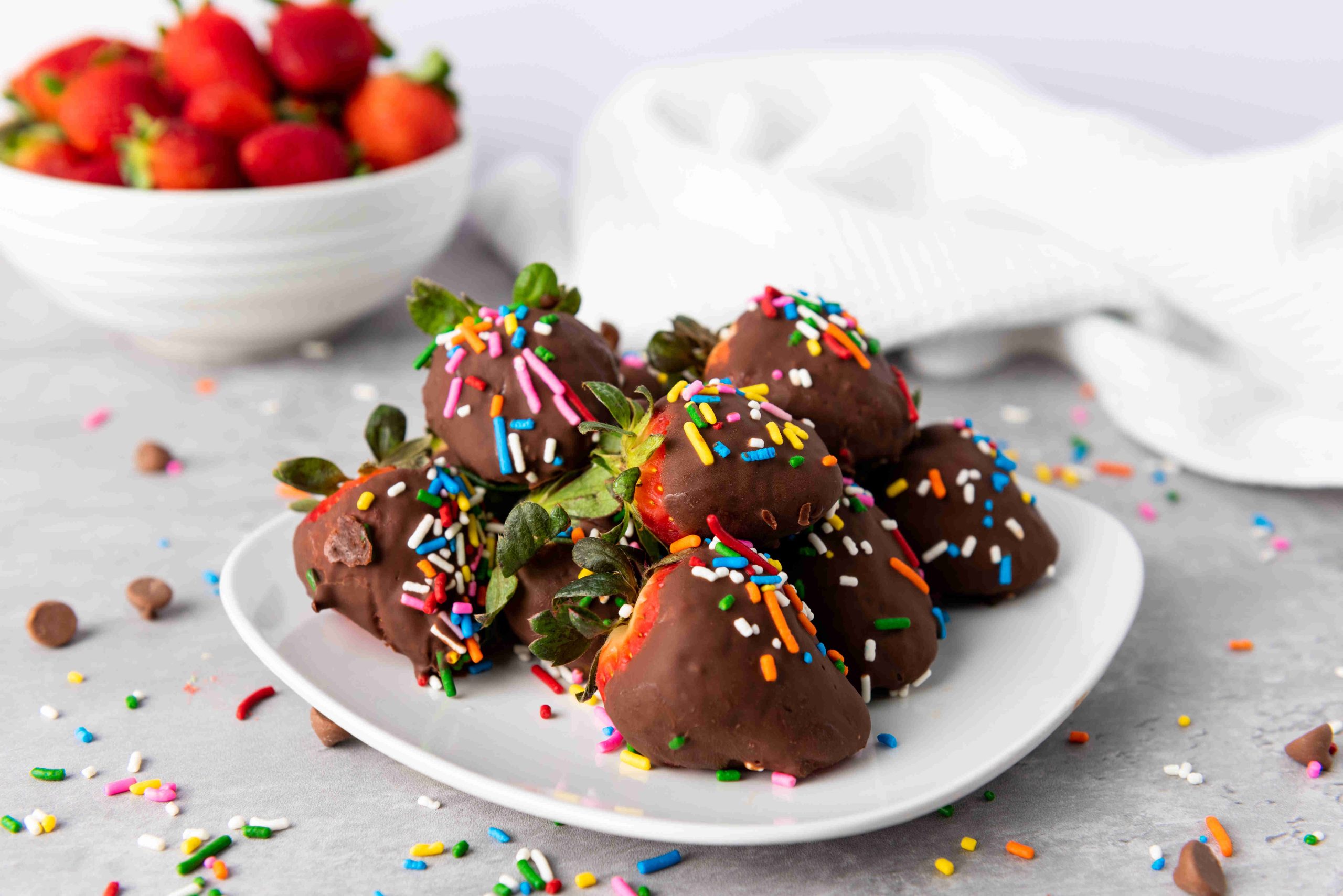 A plate of Valentine's Day Chocolate Covered Strawberries topped with colorful sprinkles sits on a light surface, with a bowl of fresh strawberries and scattered sprinkles in the background.