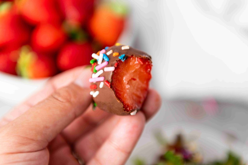 A hand holding a bitten Valentine's Day Chocolate Covered Strawberry topped with colorful sprinkles. Blurred strawberries are visible in the background.