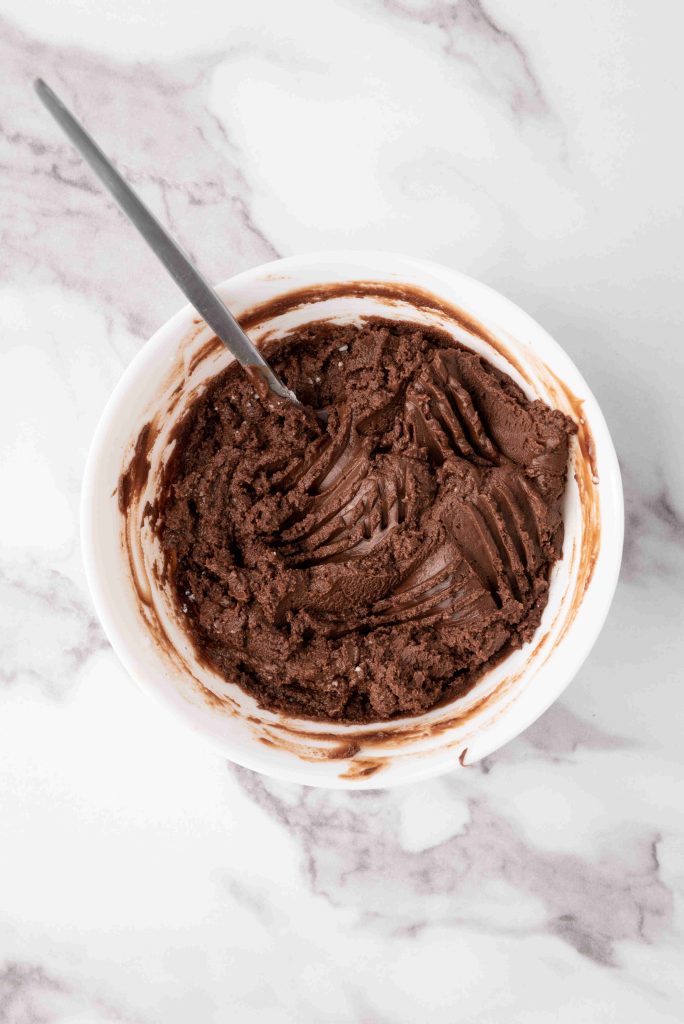 A white bowl filled with chocolate cookie dough and a metal spoon, placed on a white marble surface—perfect for making Valentine’s Day chocolate candy treats.