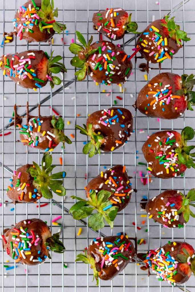 Valentine's Day Chocolate Covered Strawberries with rainbow sprinkles rest on a metal cooling rack. Some sprinkles have fallen below, and the strawberries are arranged in rows, their green leaves still attached.
