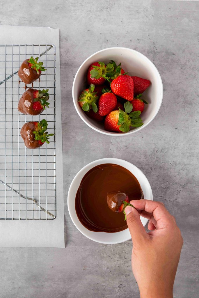 A hand dips a fresh strawberry into melted chocolate—perfect for Valentine's Day Chocolate Covered Strawberries. Nearby, a bowl of strawberries and a cooling rack with chocolate-dipped treats rest on a gray surface.