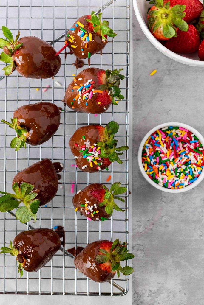 Valentine's Day Chocolate Covered Strawberries rest on a wire rack, some adorned with colorful sprinkles. Nearby, a bowl of fresh strawberries and rainbow sprinkles brighten the gray surface.