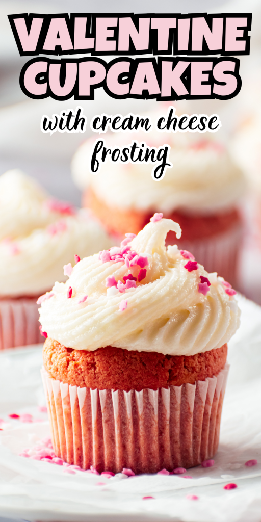 A pink Valentine's Day Cupcake with cream cheese frosting and heart-shaped sprinkles sits on a white plate. In the background, more Valentine’s Day Cupcakes can be seen. Text above reads, Valentine Cupcakes with cream cheese frosting.