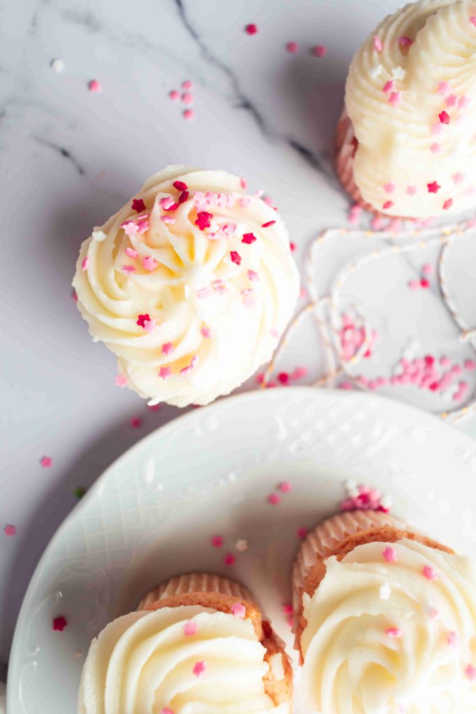 Overhead view of Valentine's Day Cupcakes with white frosting and pink and white sprinkles on a marble surface. Some cupcakes are on a white plate, while others are directly on the surface.