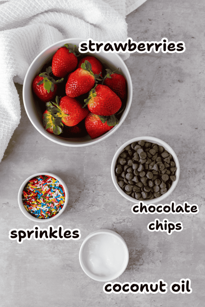 Four bowls on a gray surface hold the ingredients for Chocolate Covered Strawberries For Mothers Day: strawberries, chocolate chips, sprinkles, and coconut oil. Each is labeled in bold text. A white towel rests in the upper left corner.