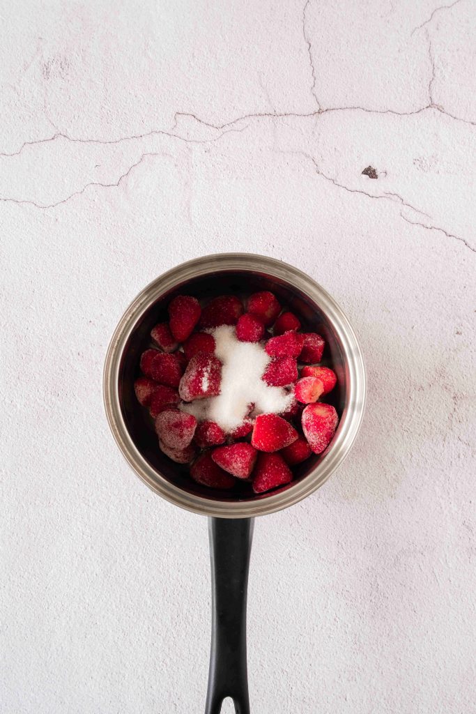 A saucepan filled with frozen strawberries and a pile of sugar sits on a textured, light-colored surface, ready to become a luscious strawberry mascarpone dessert.