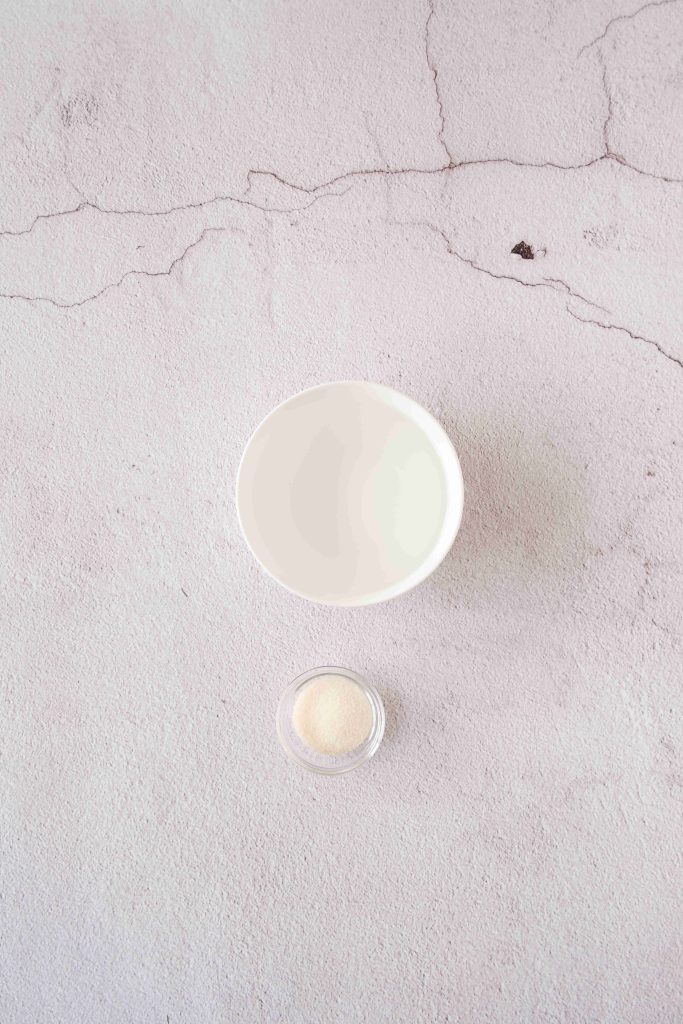 A white bowl filled with milk and a small glass dish containing gelatin powder, essential for a strawberry mascarpone dessert, are placed on a light, textured surface with subtle cracks.