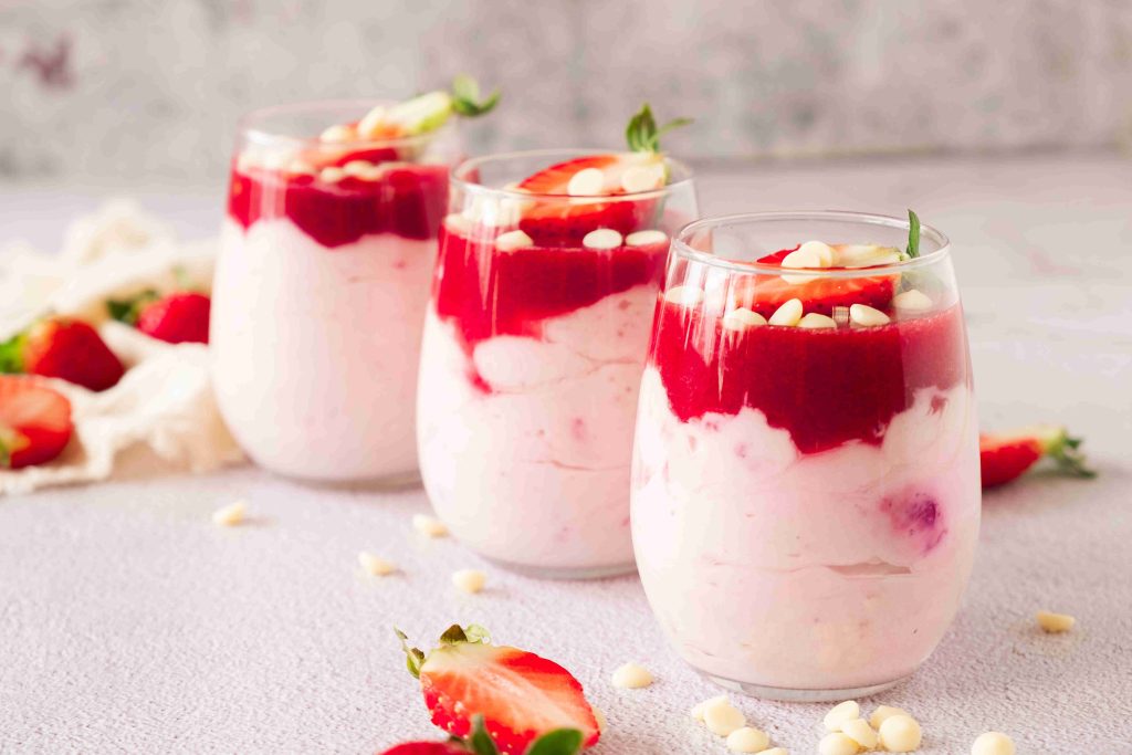 Three glasses filled with layers of strawberry mascarpone dessert, strawberry sauce, and white chocolate chips, garnished with fresh strawberry slices, sit on a light surface with scattered strawberries and chocolate chips.