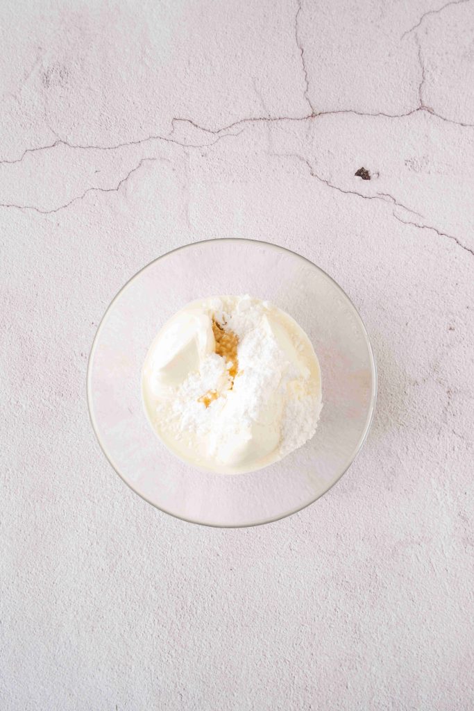 A clear glass bowl containing a mixture of white powdered sugar, cream cheese, and cream sits on a light textured surface—perfect for layering in a strawberry mascarpone dessert.