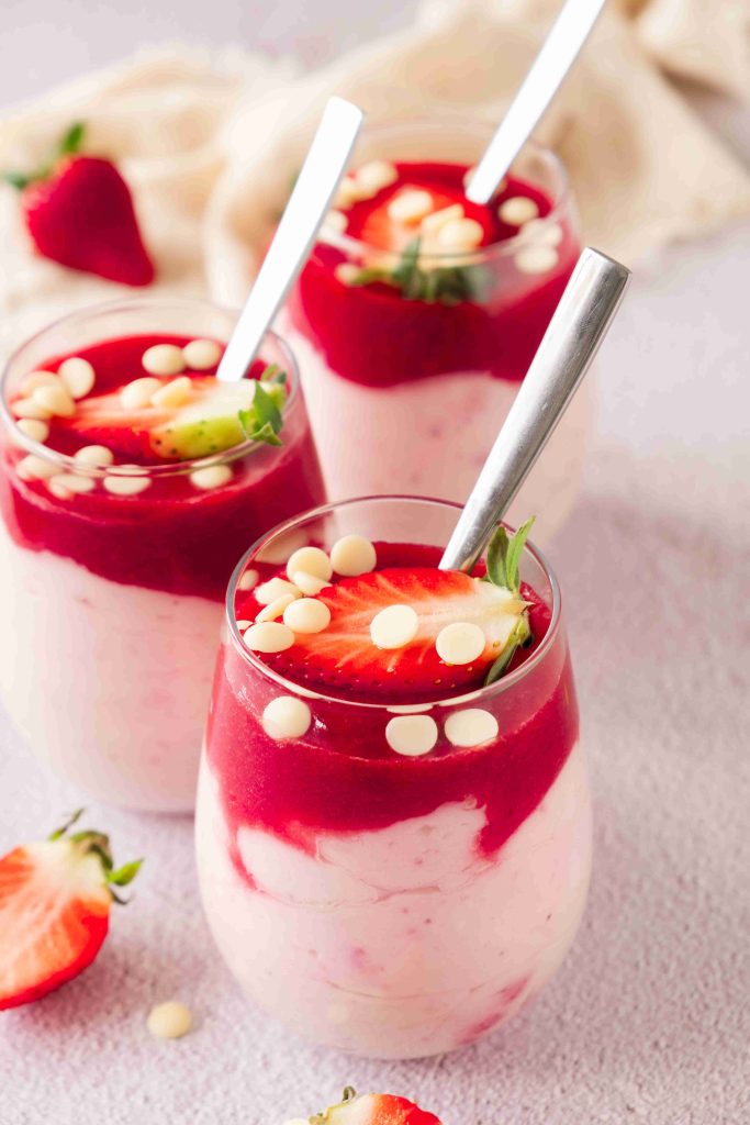 Three glasses of strawberry mascarpone dessert are topped with red strawberry sauce, fresh halved strawberries, and white chocolate chips. Each glass has a spoon and is surrounded by scattered strawberries.