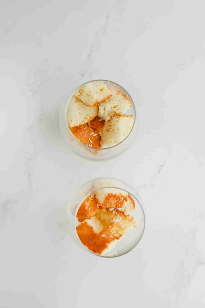 Two glass cups filled with layers of light sponge cake and orange sauce, reminiscent of a strawberry parfait with pound cake, viewed from above on a white surface.