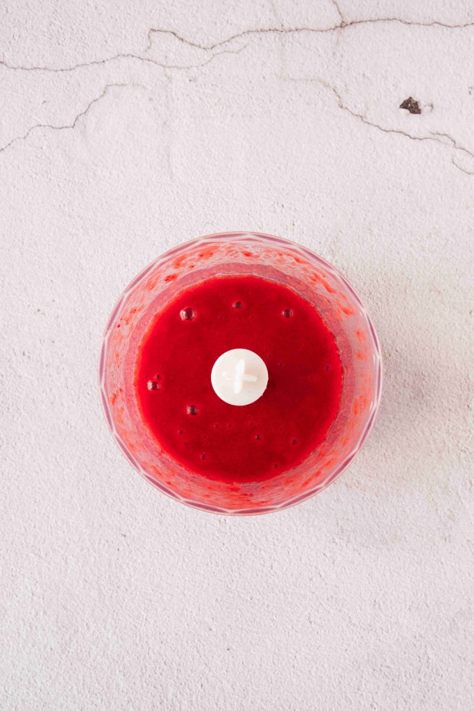 A glass blender bowl filled with bright red strawberry mascarpone dessert smoothie or sauce, viewed from above, sits on a light textured surface with visible cracks.
