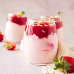 Three glasses filled with layered strawberry mascarpone dessert parfaits, topped with strawberry sauce and white chocolate chips; fresh strawberries are scattered around on a light surface.