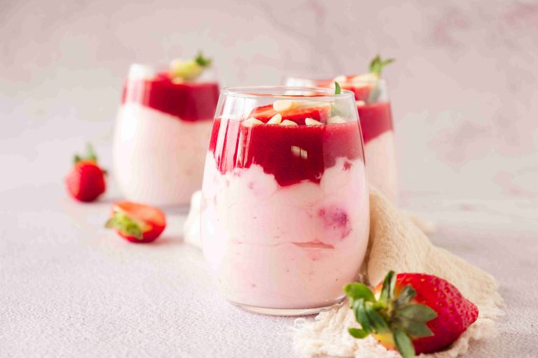 Three glasses filled with layered strawberry mascarpone dessert parfaits, topped with strawberry sauce and white chocolate chips; fresh strawberries are scattered around on a light surface.