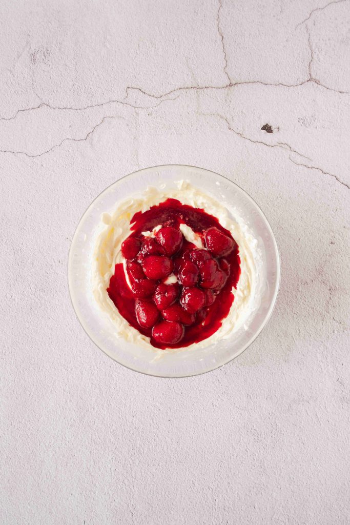 A glass bowl filled with whipped cream and topped with cherry compote sits on a light, textured surface, creating an inviting strawberry mascarpone dessert.