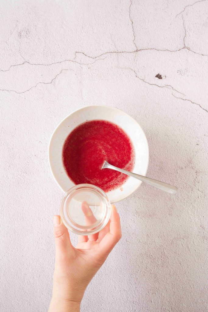 A hand holds a small empty glass bowl above a larger white bowl filled with a red liquid mixture for a strawberry mascarpone dessert, with a spoon resting inside, on a light textured surface.
