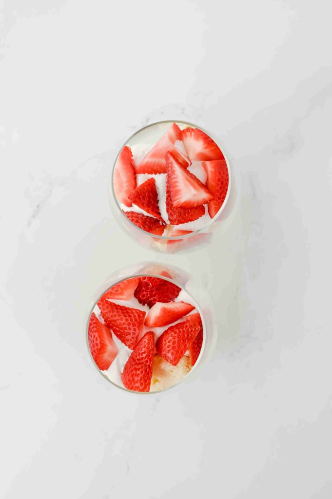 Two clear glasses filled with a creamy strawberry parfait with pound cake, topped with sliced fresh strawberries, viewed from above on a white surface.