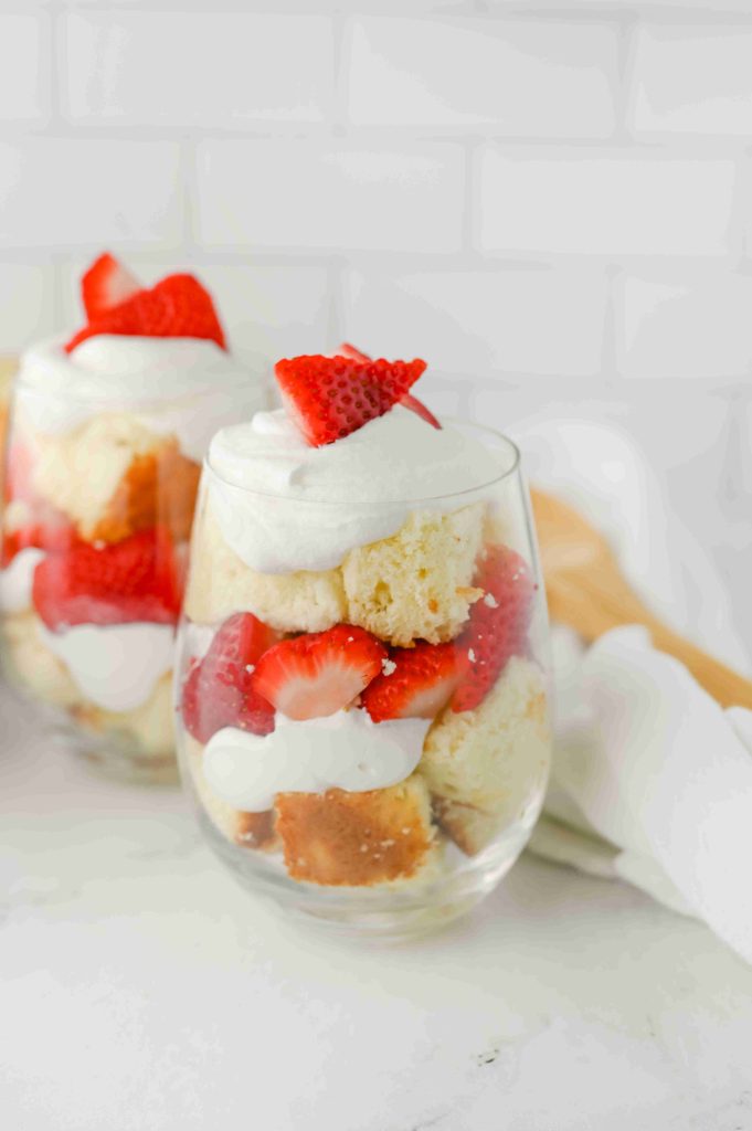 Two clear glasses filled with layers of strawberry parfait with pound cake, whipped cream, and sliced strawberries, topped with a dollop of whipped cream and a strawberry slice, set on a white surface.