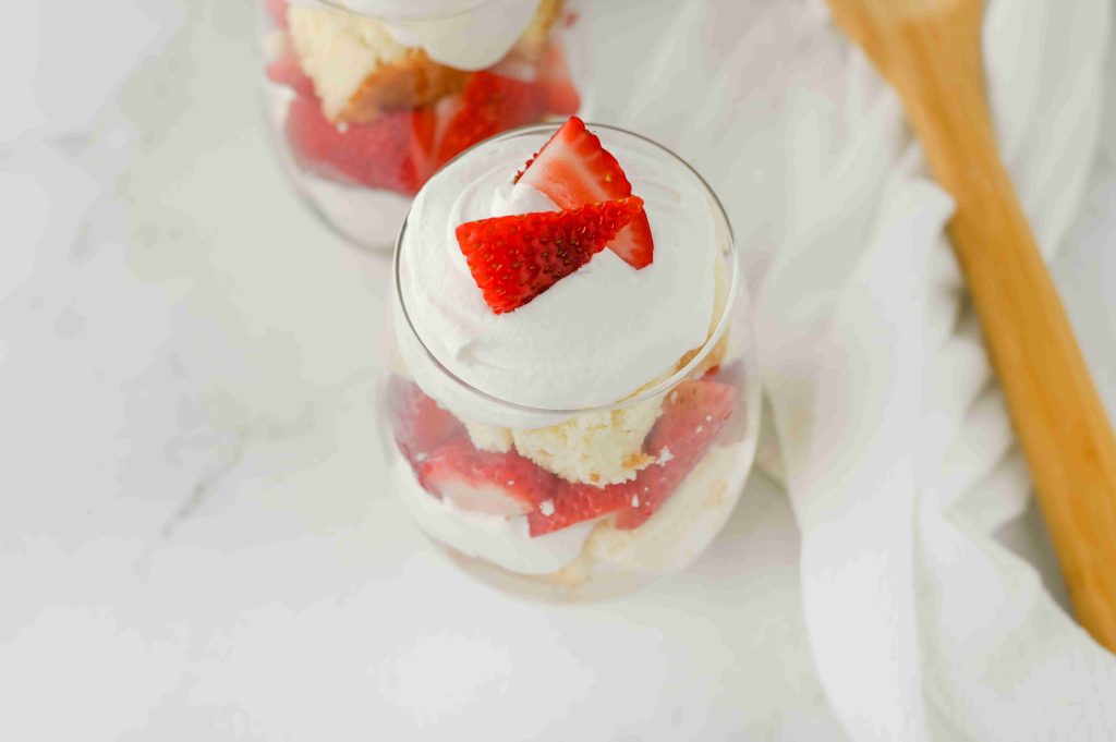 A glass dessert cup filled with layers of whipped cream, sliced strawberries, and pound cake creates a delightful strawberry parfait with pound cake, topped with two strawberry slices. A wooden spoon and white cloth are nearby on a white surface.