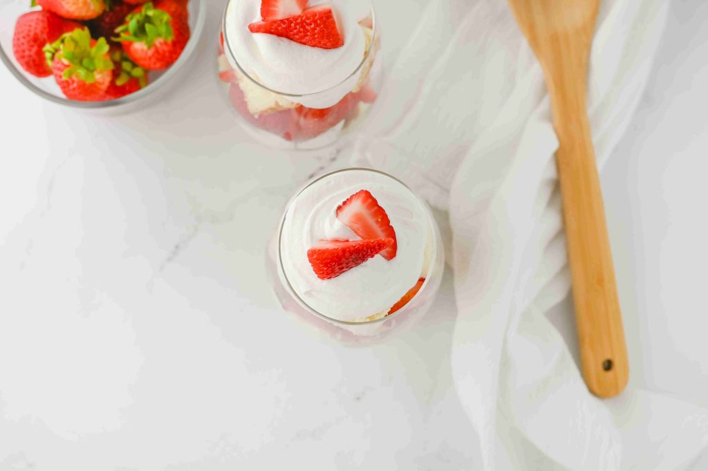 Overhead view of two strawberry parfait with pound cake glasses topped with whipped cream and sliced strawberries, next to a bowl of whole strawberries, a wooden spoon, and a white cloth on a light surface.