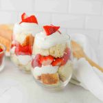 Two glasses filled with layers of strawberry parfait with pound cake, whipped cream, and sliced strawberries, topped with a strawberry fan. A bowl of whole strawberries and a wooden spoon are in the background on a white countertop.
