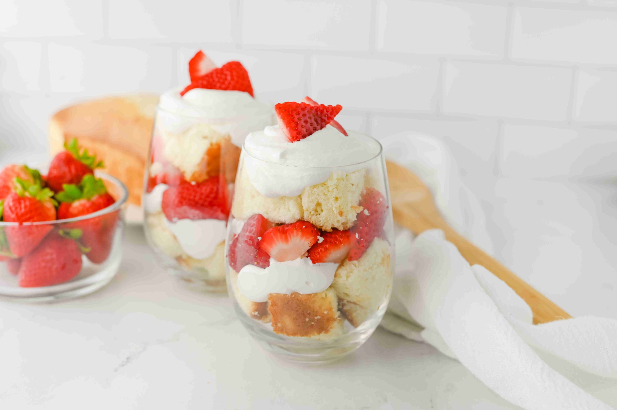 Two glasses filled with layers of strawberry parfait with pound cake, whipped cream, and sliced strawberries, topped with a strawberry fan. A bowl of whole strawberries and a wooden spoon are in the background on a white countertop.