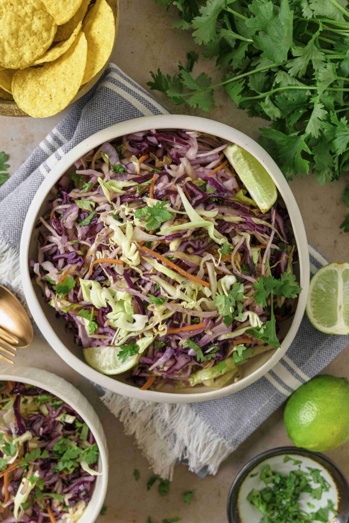 A bowl of colorful Mexican slaw no mayo with cilantro and lime wedges sits on a striped napkin, surrounded by fresh cilantro, lime, tortilla chips, and a small bowl of extra slaw.