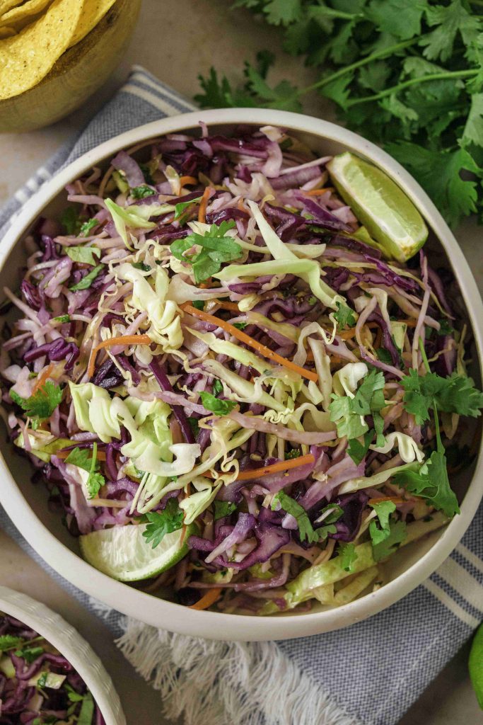 A bowl of colorful Mexican slaw no mayo made with shredded green and purple cabbage, carrots, cilantro, and lime wedges sits on a gray napkin with tortilla chips and fresh cilantro nearby.
