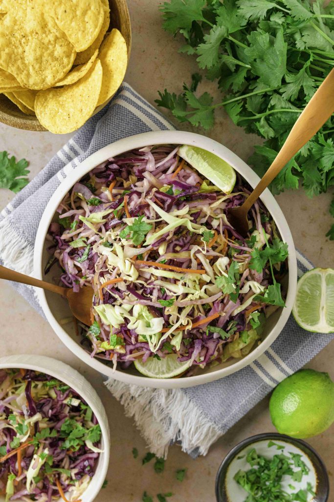 A bowl of colorful Mexican slaw no mayo with cilantro, served with lime wedges and tortilla chips on the side. Fresh cilantro and a bowl of chips rest nearby on a striped cloth.