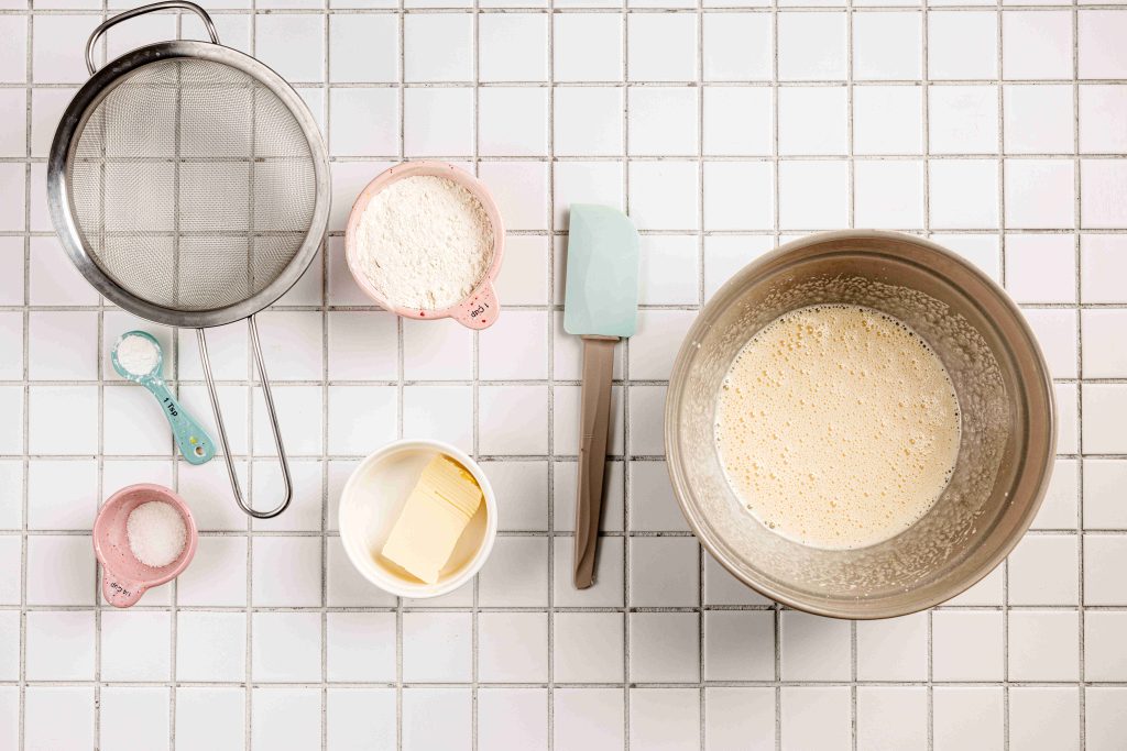 Baking ingredients on a tiled surface, including flour, sugar, butter, a whisked mixture in a bowl, and tools—perfect for creating delicious treats like a strawberry mascarpone trifle.