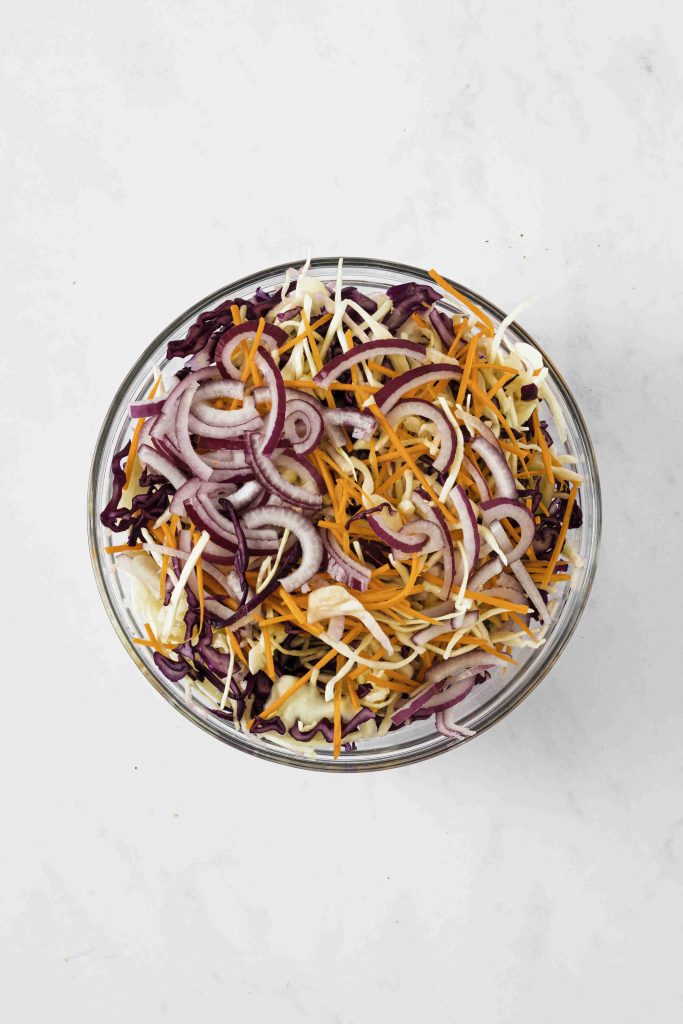 A glass bowl filled with shredded purple cabbage, orange carrots, and sliced red onions creates a vibrant Mexican slaw no mayo, all mixed together on a white marble surface.