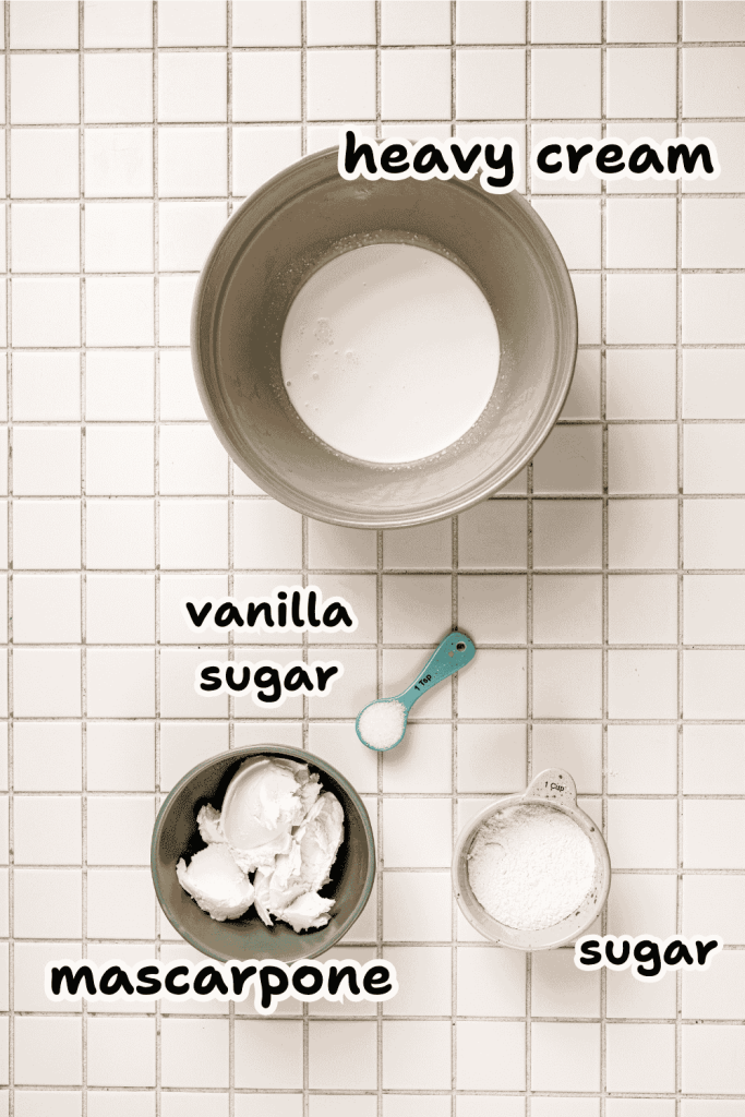 Top-down view of bowls with labeled ingredients on a tiled surface: heavy cream, mascarpone, sugar, and a teaspoon of vanilla sugar—perfect for assembling a delicious strawberry mascarpone trifle.