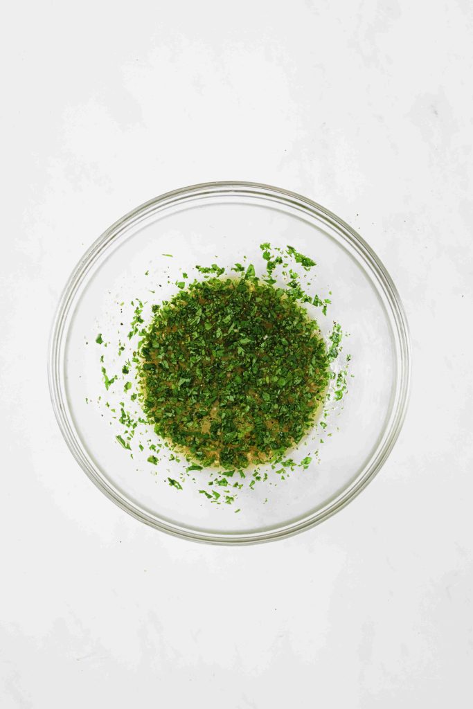 A clear glass bowl containing a mixture of finely chopped green herbs and seasonings for mexican slaw no mayo, placed on a white surface.