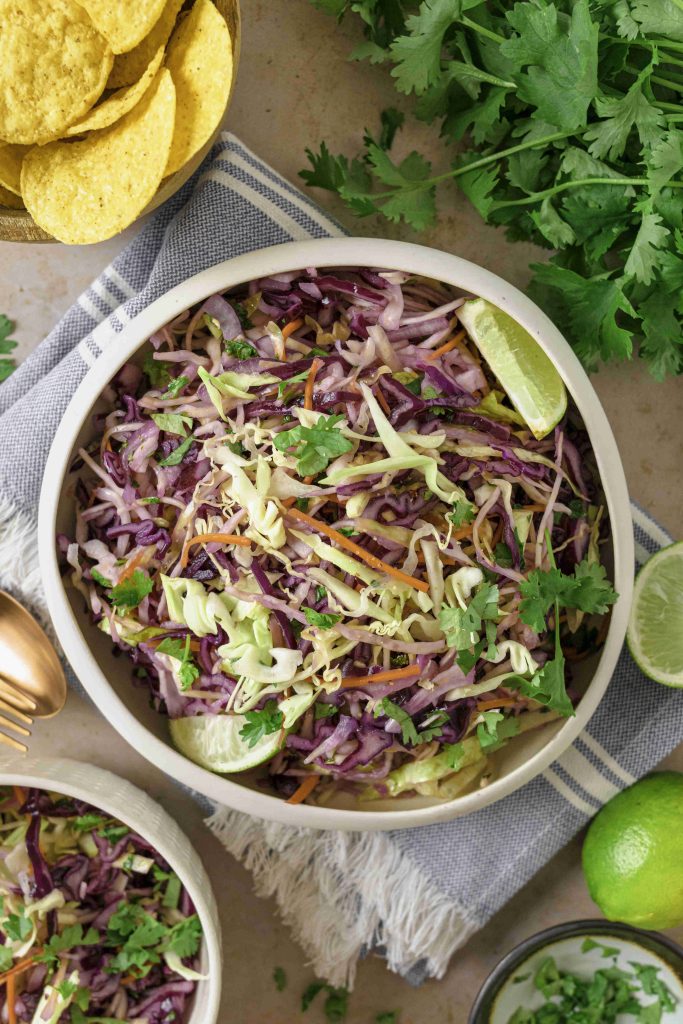A bowl of colorful Mexican slaw no mayo with cilantro and lime wedges sits on a striped cloth next to tortilla chips, fresh cilantro, and limes. A fork and another bowl of vibrant slaw are also visible.