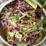 A bowl of vibrant Mexican slaw no mayo with shredded green and purple cabbage, carrots, and fresh cilantro, garnished with lime wedges. Two gold utensils rest in the bowl atop a striped napkin underneath.