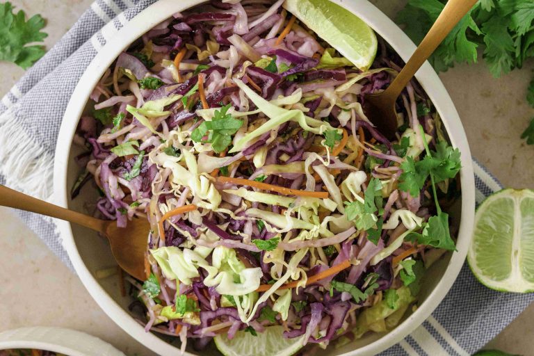 A bowl of vibrant Mexican slaw no mayo with shredded green and purple cabbage, carrots, and fresh cilantro, garnished with lime wedges. Two gold utensils rest in the bowl atop a striped napkin underneath.