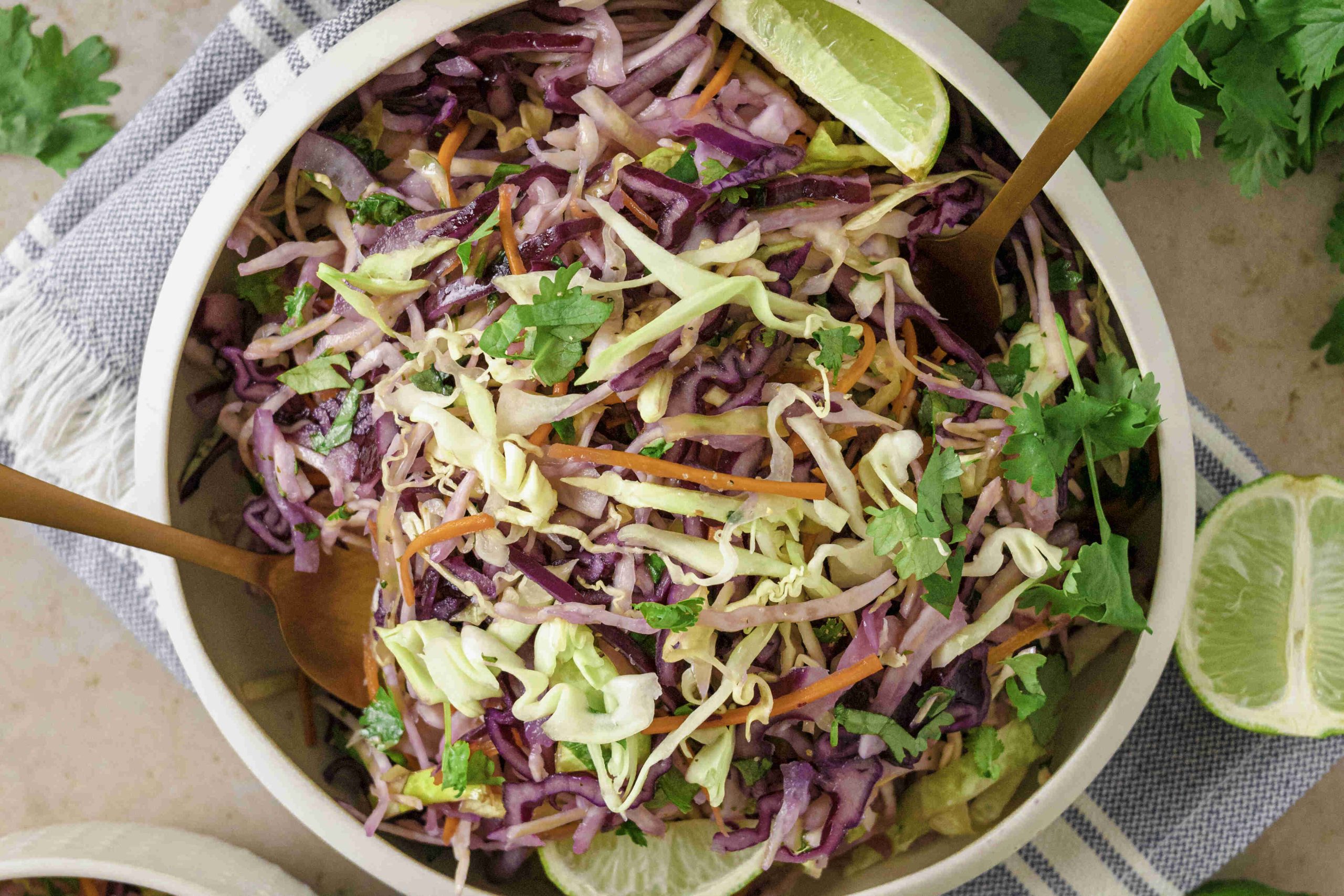 A bowl of vibrant Mexican slaw no mayo with shredded green and purple cabbage, carrots, and fresh cilantro, garnished with lime wedges. Two gold utensils rest in the bowl atop a striped napkin underneath.