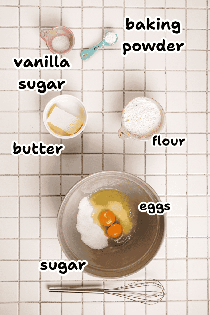 Top-down view of baking ingredients on a tiled surface for a strawberry mascarpone trifle: labeled bowls of baking powder, vanilla sugar, flour, butter, eggs, and sugar, with a whisk beside a mixing bowl containing eggs and sugar.