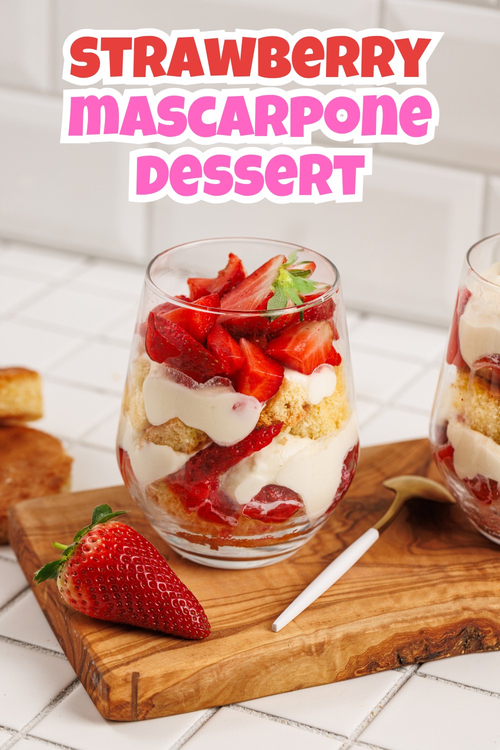 A glass filled with layers of strawberries, mascarpone cream, and cake sits on a wooden board next to a fresh strawberry and a gold spoon, showcasing a delicious strawberry mascarpone trifle. The text above reads “Strawberry Mascarpone Dessert.”.