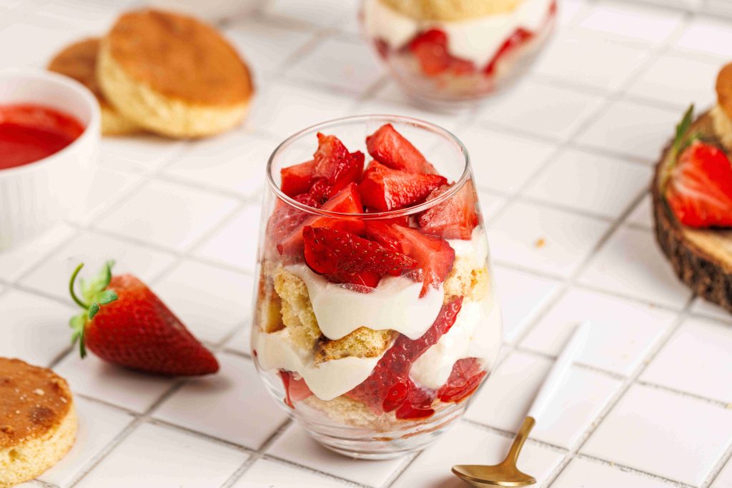 A glass filled with layers of whipped cream, sliced strawberries, and biscuit pieces sits on a white tiled surface, surrounded by fresh strawberries, biscuits, and a gold spoon—perfect for a strawberry mascarpone trifle.
