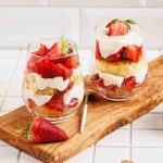 Two glass cups filled with layers of sliced strawberries, whipped cream, cake, and creamy mascarpone make a tempting strawberry mascarpone trifle on a wooden board. A fresh strawberry sits nearby, with dessert ingredients in the tiled background.
