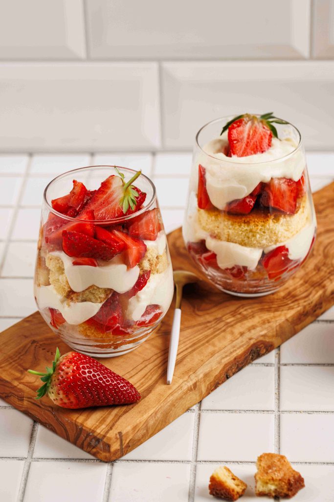 Two glasses filled with layers of strawberries, cream, and sponge cake create a tempting strawberry mascarpone trifle on a wooden board, with a fresh strawberry and cake crumbs nearby against a white tiled background.