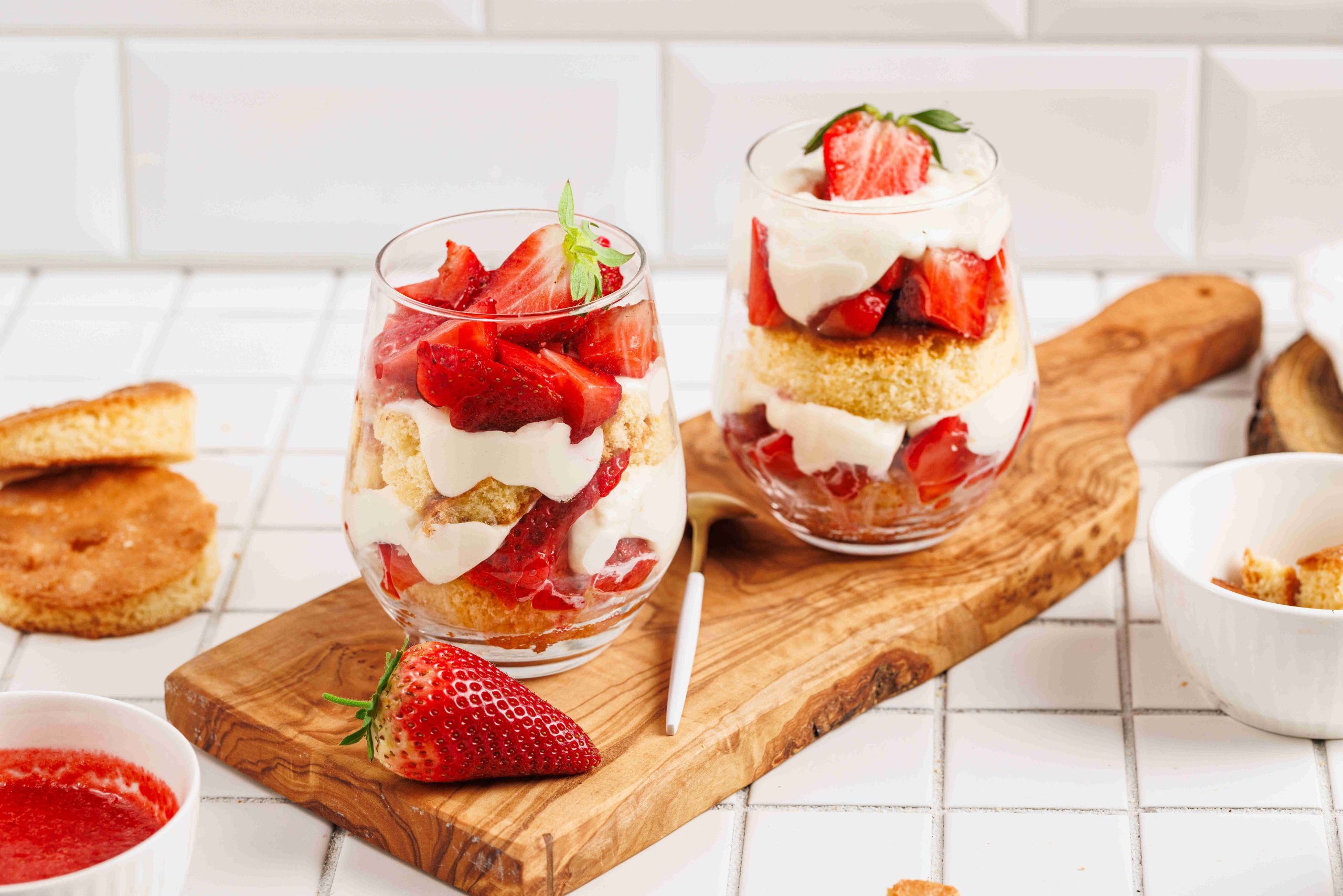 Two glass cups filled with layers of sliced strawberries, whipped cream, cake, and creamy mascarpone make a tempting strawberry mascarpone trifle on a wooden board. A fresh strawberry sits nearby, with dessert ingredients in the tiled background.