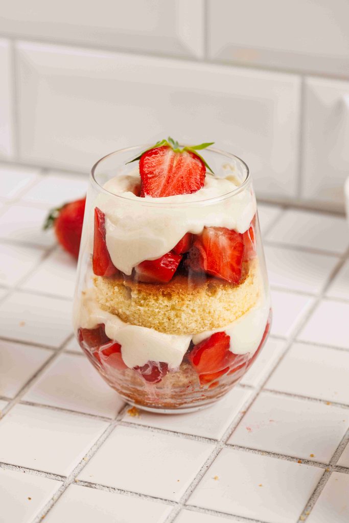 A dessert glass filled with layers of sliced strawberries, sponge cake, and creamy whipped topping creates a delectable strawberry mascarpone trifle, garnished with a halved strawberry on top and set on a white tiled surface.