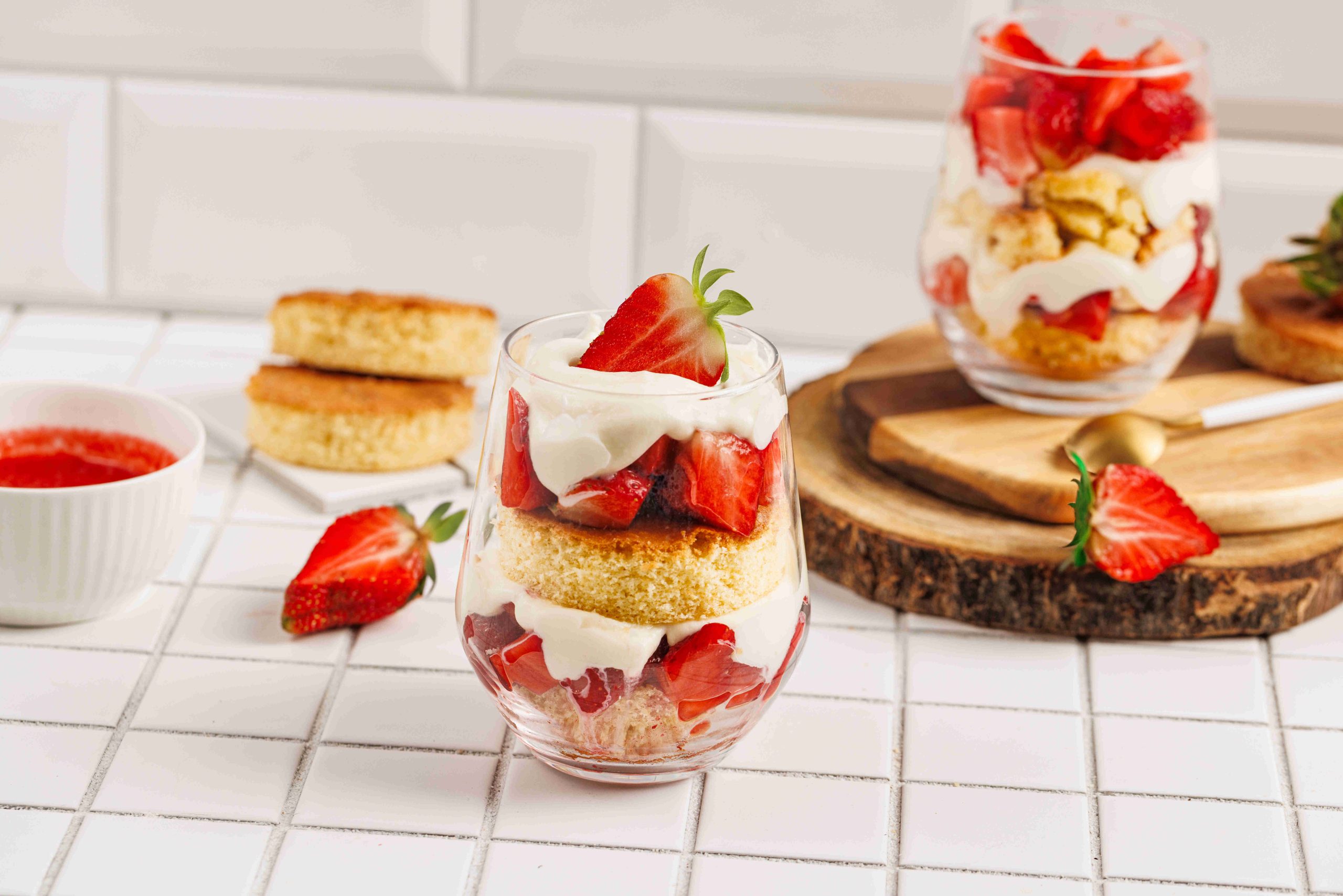 A glass filled with layers of sponge cake, whipped cream, and fresh strawberries creates a tempting strawberry mascarpone trifle on a tiled surface, surrounded by more strawberry desserts and cake pieces in the background.