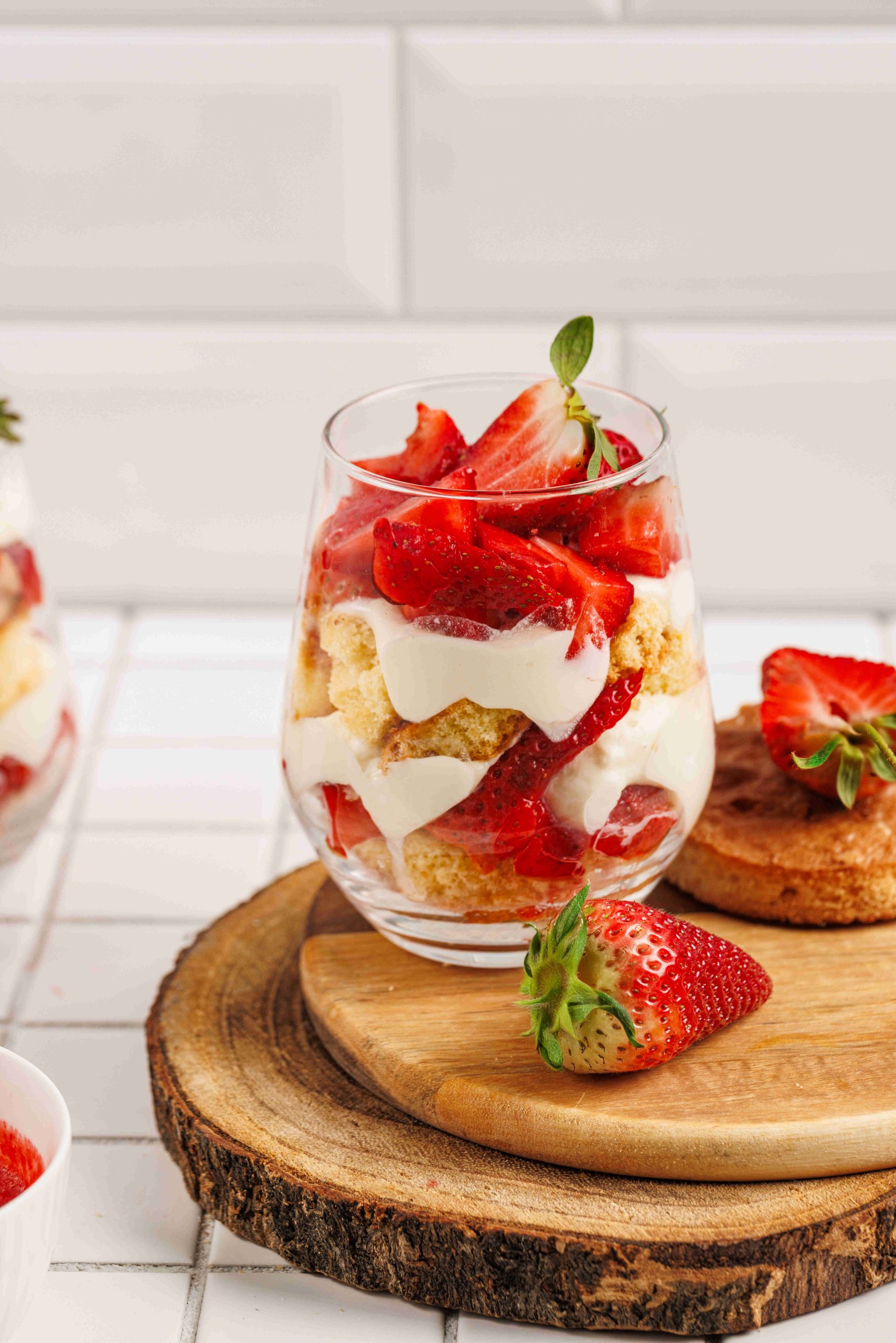 A glass filled with layers of whipped cream, sponge cake, and fresh strawberries sits on a wooden board—an inviting strawberry mascarpone trifle. A whole strawberry and a piece of cake rest nearby, with another dessert glass in the background.