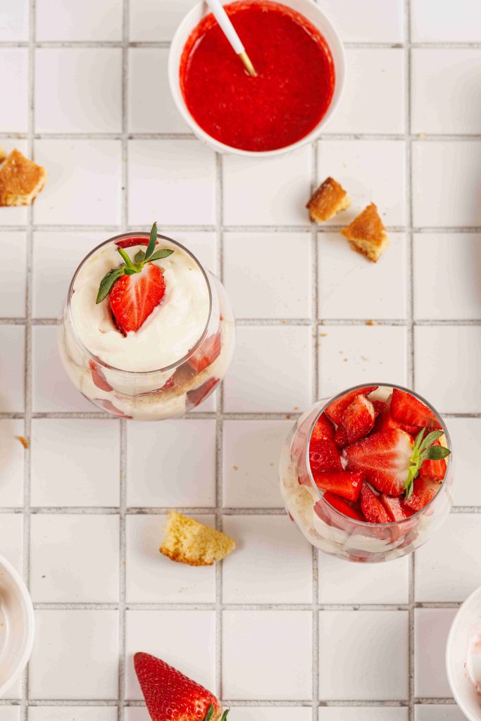 Two glasses of strawberry mascarpone trifle topped with fresh strawberries are on a tiled surface, surrounded by a bowl of strawberry sauce, cake crumbs, and a whole strawberry.